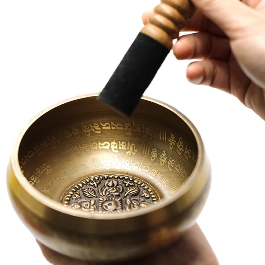 A person holding a mallet above a bronze-colored hand-hammered Tibetan singing bowl with inscriptions and a floral design inside.
